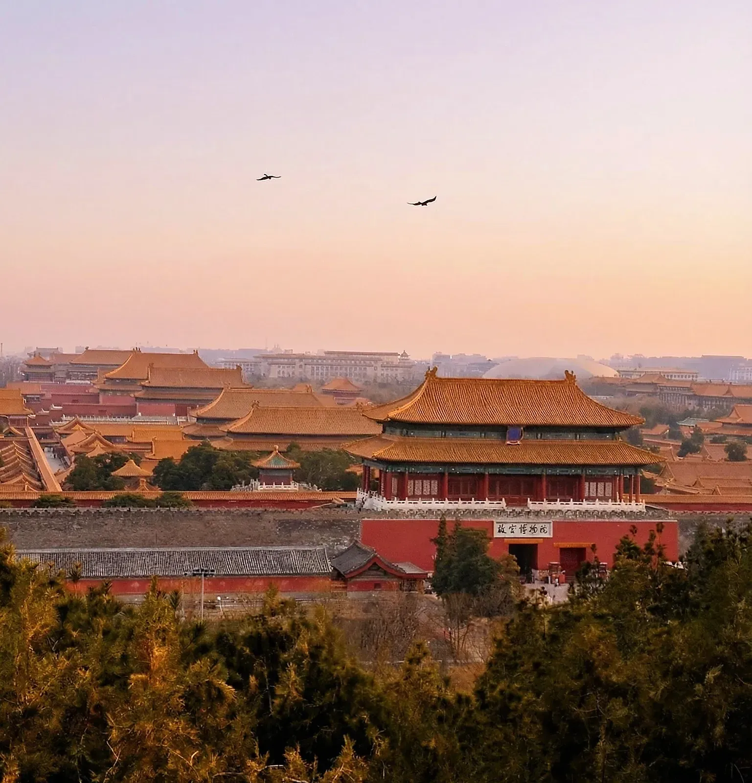 Golden palace roofs behind a long red wall at hazy dusk.