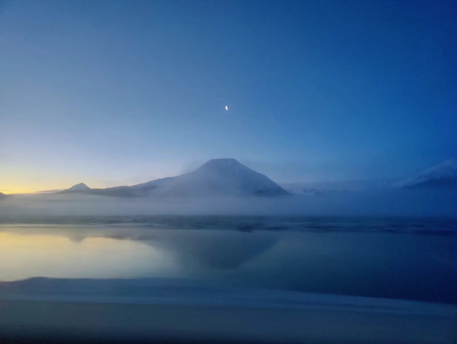 Crescent moon above a mist-wrapped peak reflected in water.